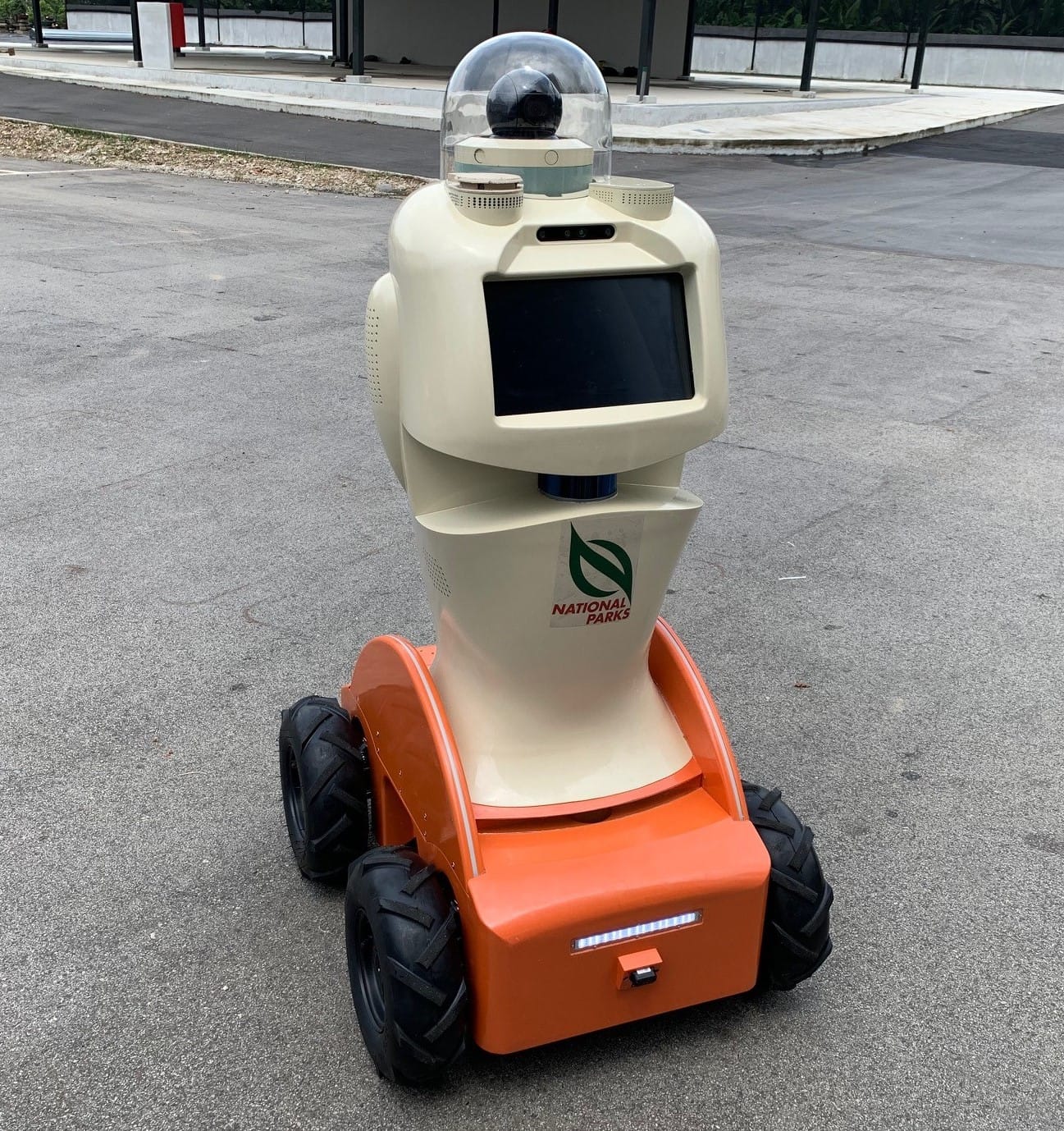 A four-wheeled robot with an orange base, beige body, screen, and camera labeled "National Parks" on a concrete path.